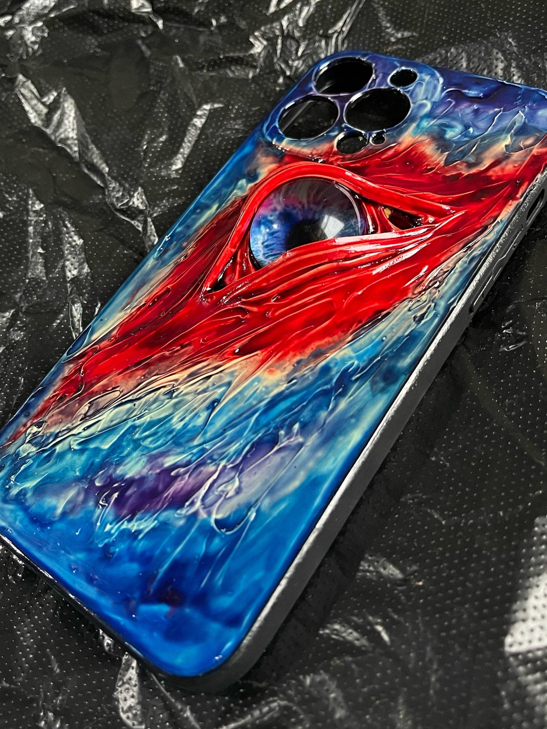 A 3D handmade iPhone case featuring a realistic blue eye design with red and blue textured cream glue accents.