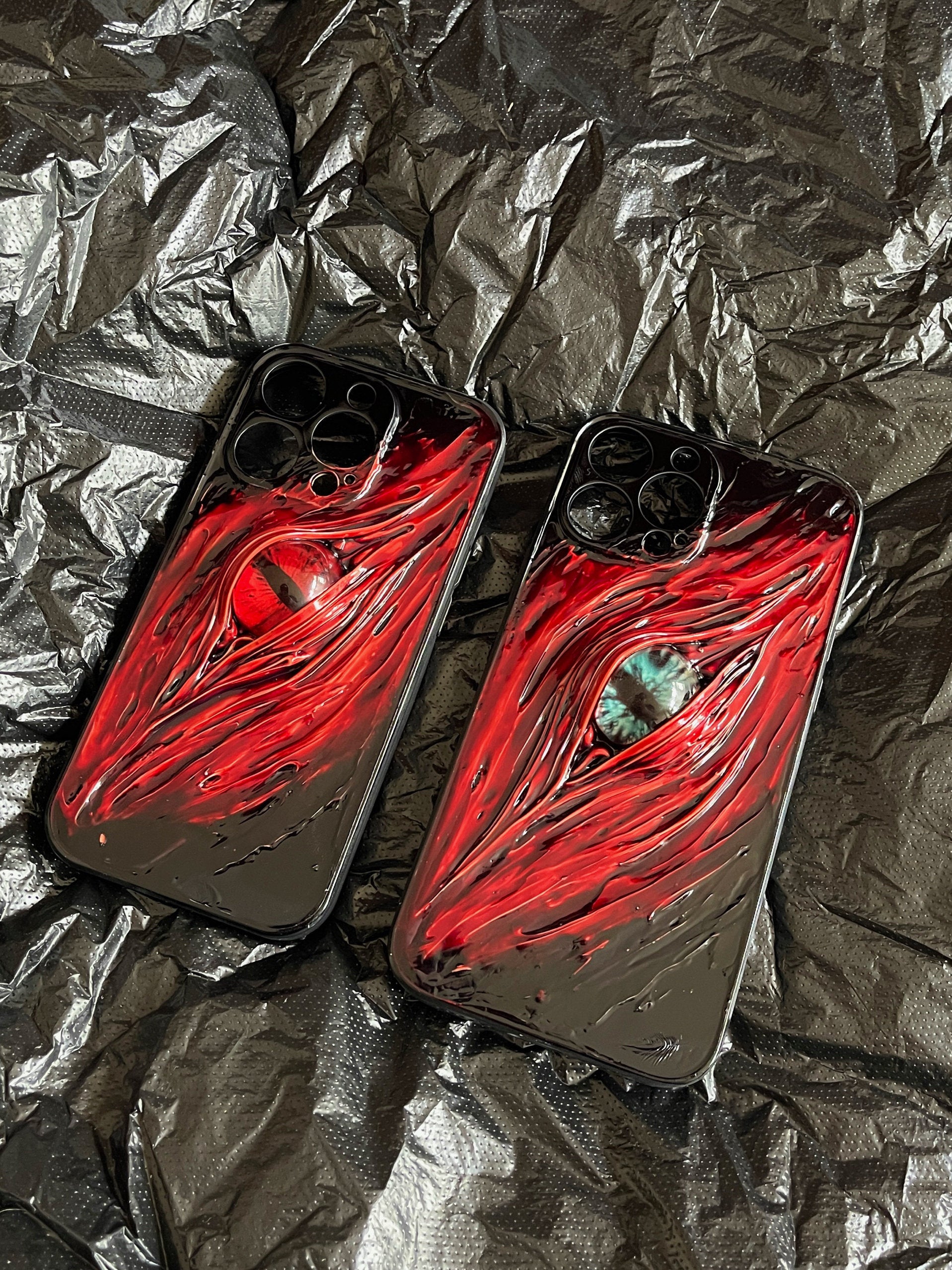 A 3D dark-style iPhone case featuring a realistic, glowing red eye design with textured, blood-like details on a black background.