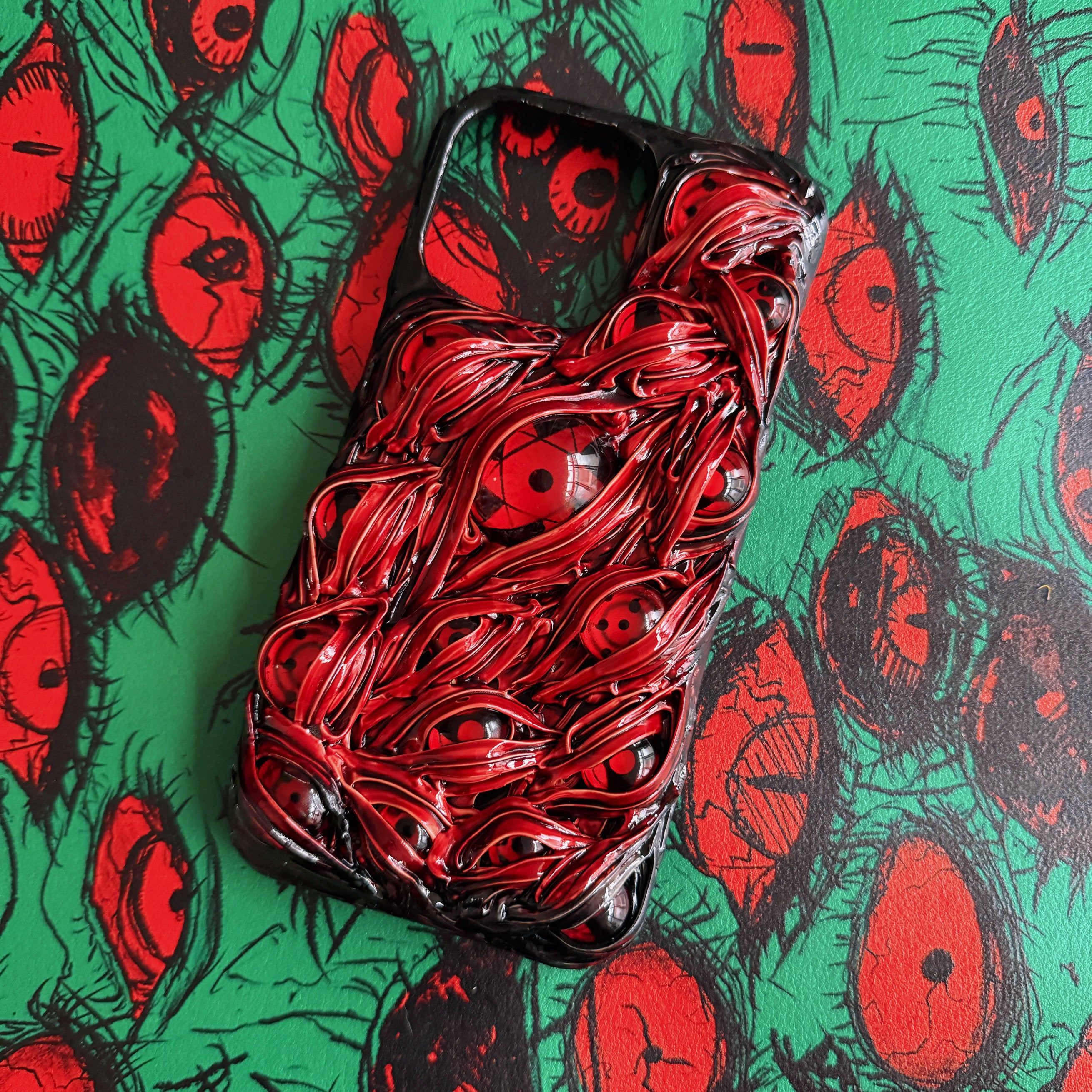 Two 3D textured anime-style iPhone cases featuring red Sharingan eyes on black and red backgrounds.