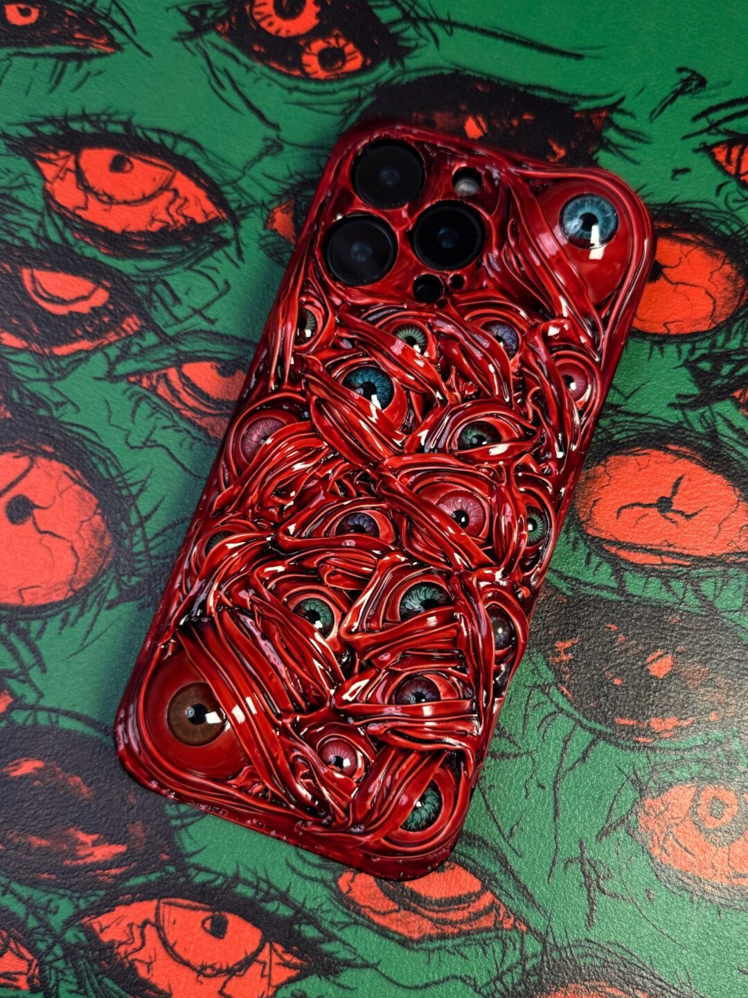 Four 3D Cthulhu-style horror eye iPhone cases in green, black, red, and purple arranged on a matching eye-patterned background.