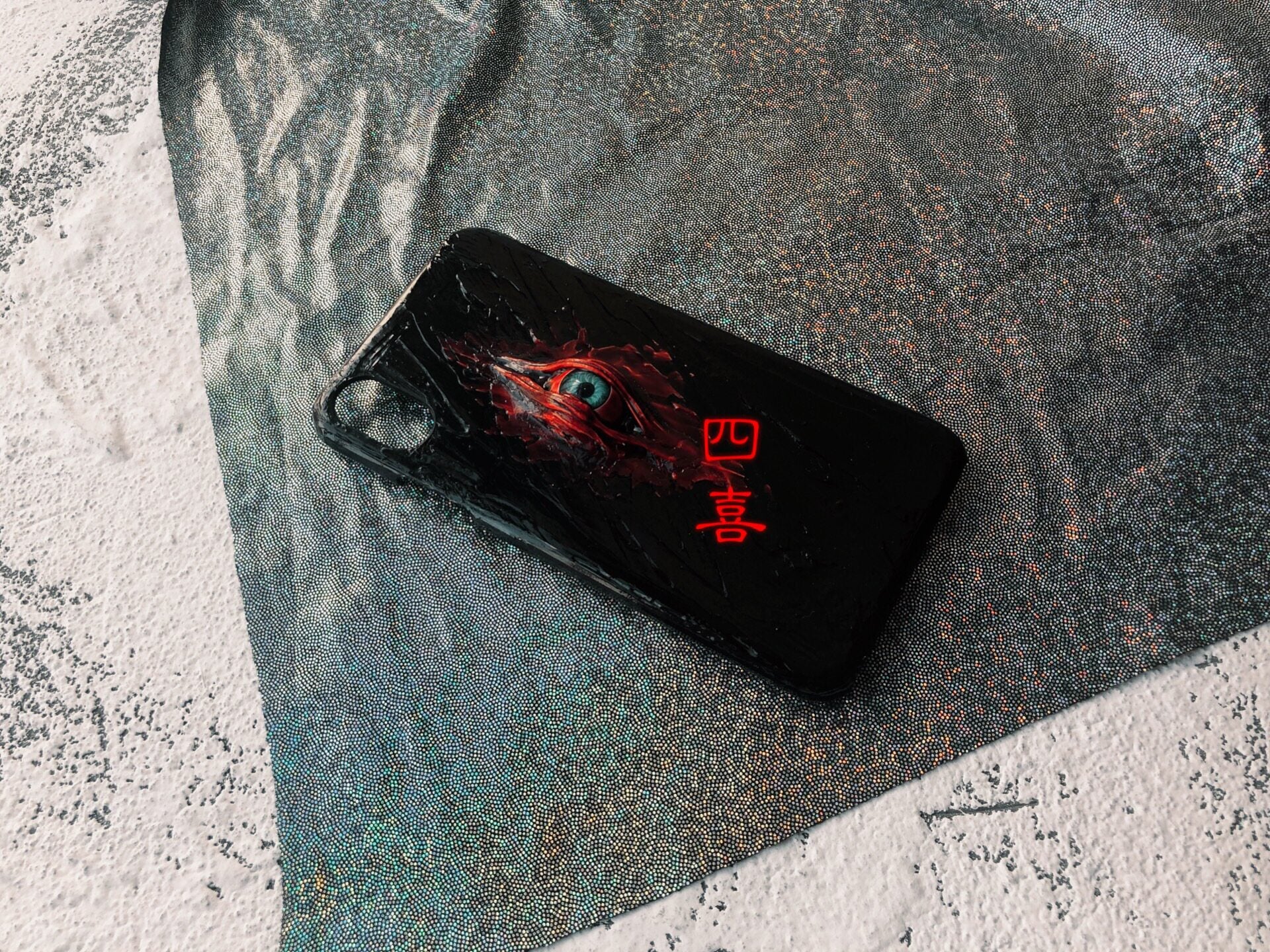 A black iPhone case featuring a realistic, creepy blue eye surrounded by red, textured blood-like details.